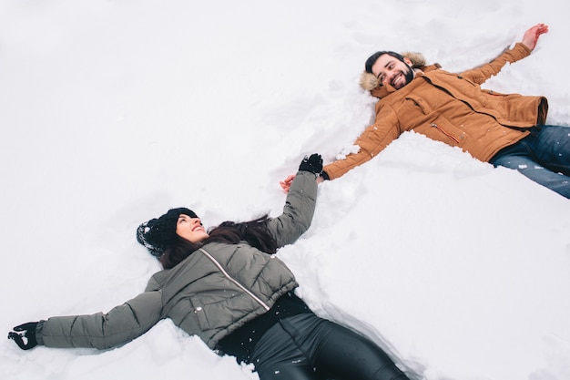 Happy Young Couple in Winter . Family Outdoors. man and woman looking upwards and laughing. Love, fun, season and people - walking in winter park. Lying in fresh snow, making snow angels having fun