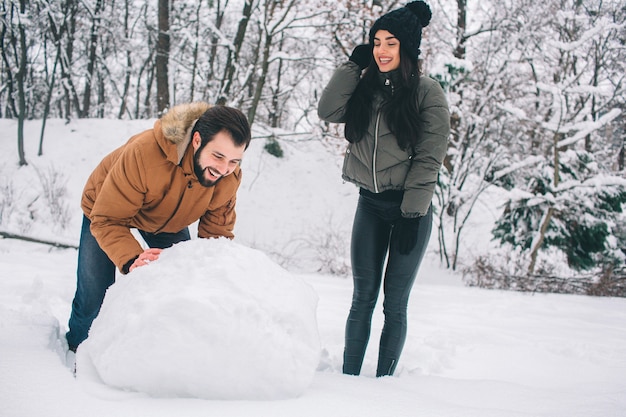 Happy Young Couple in Winter . Family Outdoors. man and woman looking upwards and laughing. Love, fun, season and people - walking in winter park. Making a snowman.