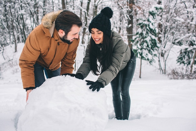 Happy Young Couple in Winter . Family Outdoors. man and woman looking upwards and laughing. Love, fun, season and people - walking in winter park. Making a snowman.