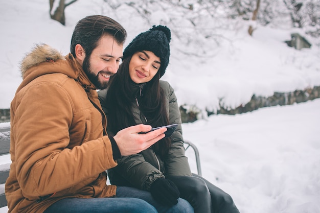 Happy Young Couple in Winter . Family Outdoors. man and woman looking upwards and laughing. Love, fun, season and people - walking in winter park. Sit on the bench. Holds a smartphone.