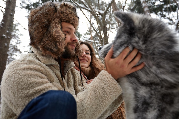 Happy young couple in winterwear playing with purebred siberian husky and having fun in snowbound pine forest