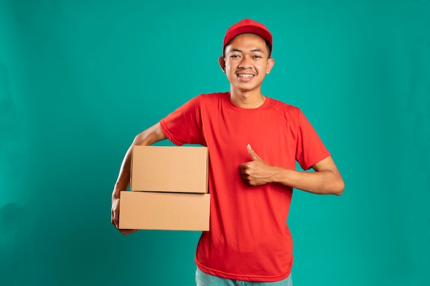 Happy young courier holding cardboard box and smiling while giving thumbs up on green background