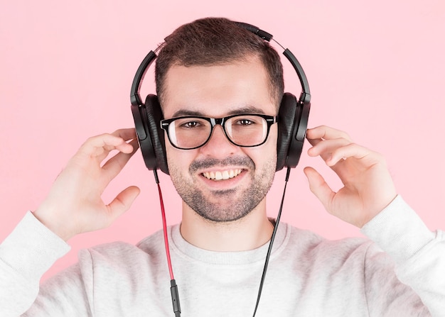 Happy young cute guy listens to music in big white headphones on a pink background, holds them, in a white sweatshirt, with a smile