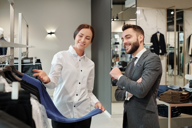 Happy young elegant man in grey suit consulting with shop assistant while looking for new pullover in boutique