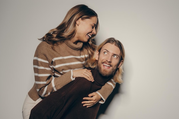 Happy young elegant man piggybacking his beautiful girlfriend while standing on beige background