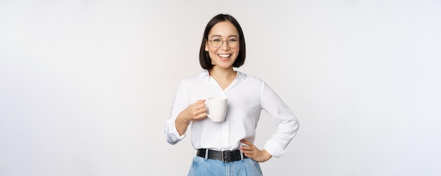 Happy young energetic asian woman smiling drinking holding cup mug of coffee standing confident against white background