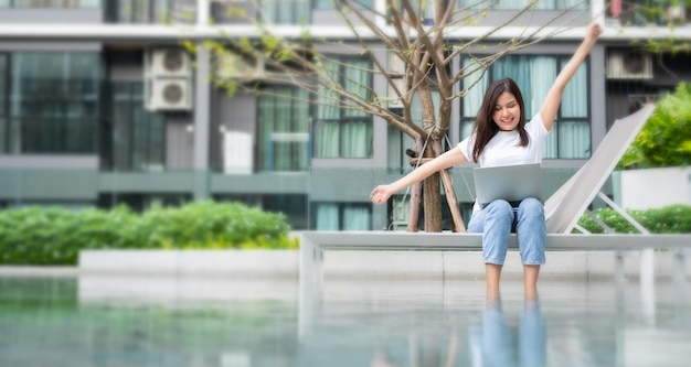 Happy young entrepreneur woman sitting on tanning bed beside pool and using laptop computer for remote online working digital online business project in quiet yard of resort house Work on vacation