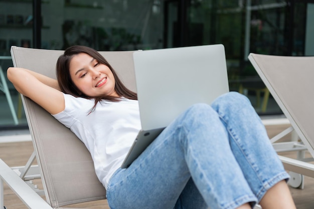 Happy young entrepreneur woman sitting on tanning bed beside pool and using laptop computer