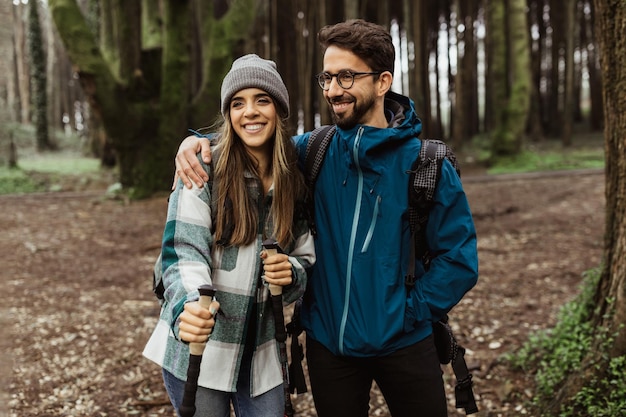 Happy young european male and woman tourists in jackets with backpack and trekking sticks walk