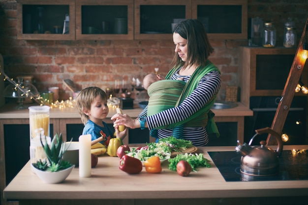 Happy young family, beautiful mother with two children, adorable preschool boy and baby in sling cooking together in a sunny kitchen.