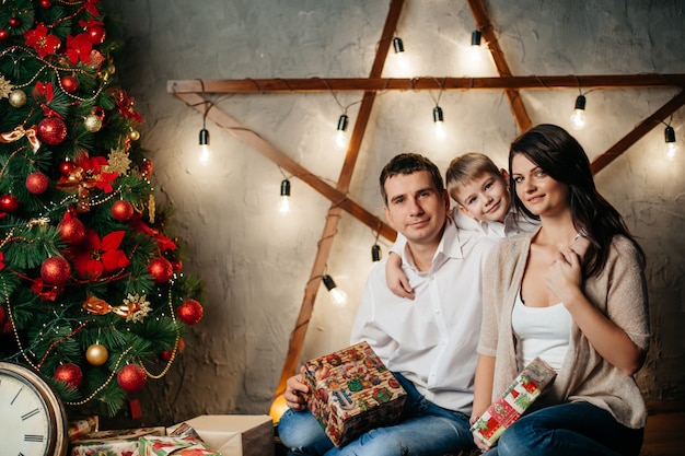 Happy young family in Christmas decorations, mom, dad and little boy near Christmas tree with presents near