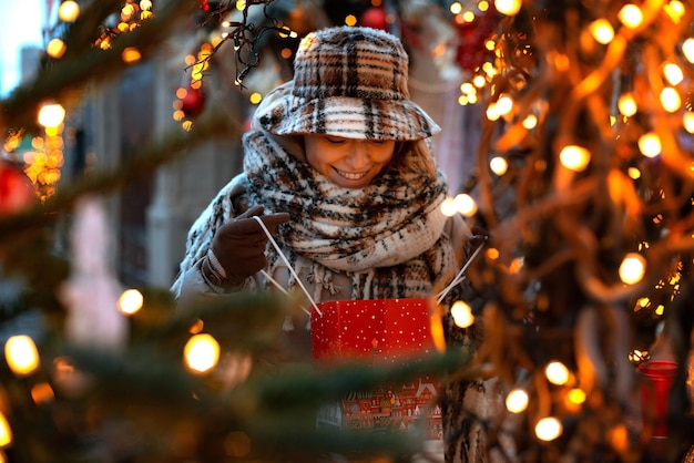 Happy young fashionable woman in a hat looks into a bag with a purchased gift at a christmas market ...