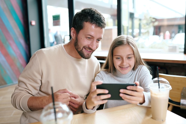 Happy young father and his daughter watching funny video in smartphone while spending time in cafe