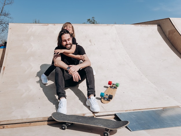 Happy young father and his son dressed in the stylish casual clothes are sitting in an embrace together on the slide next to the skateboards in a skate park at the sunny warm day .