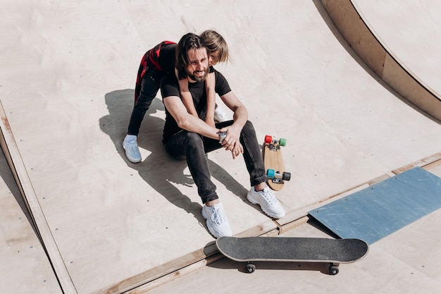 Happy young father and his son dressed in the stylish casual clothes are sitting in an embrace together on the slide next to the skateboards in a skate park at the sunny warm day .