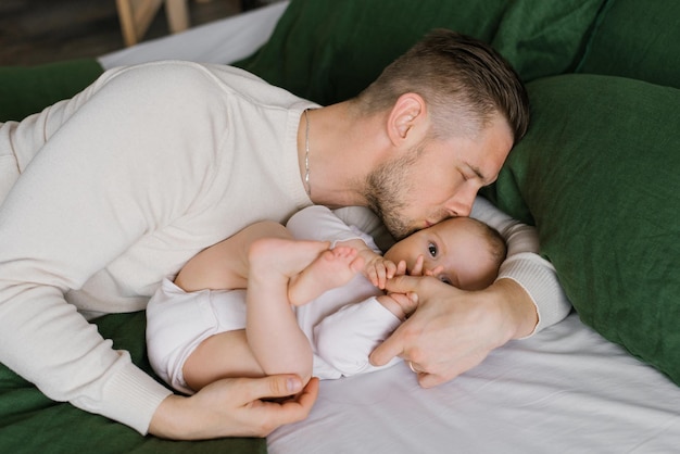 A happy young father lies on the bed with his sixmonthold son on the bed and kisses him