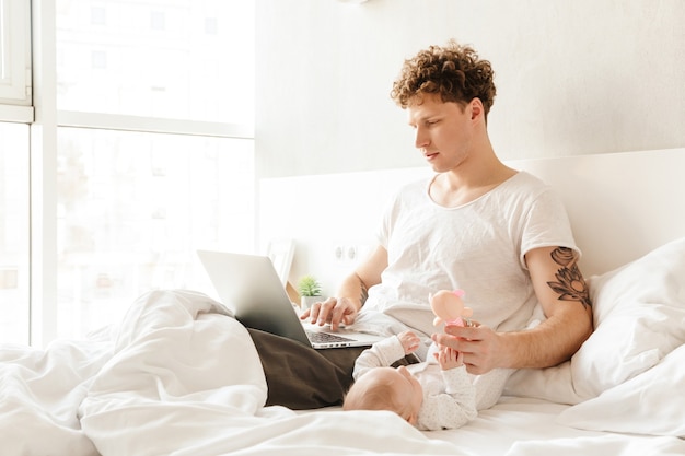 Happy young father playing with his little baby son while working on a laptop computer, laying in bed