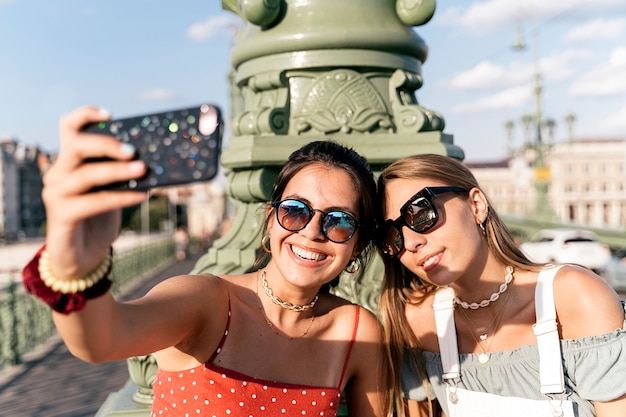 Happy young female friends in sunglasses sitting near column and browsing internet on mobile phone  of city street