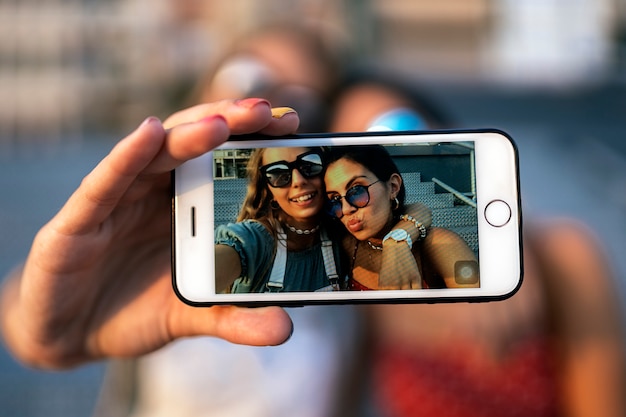 Happy young female friends in sunglasses sitting and taking a selfie on mobile phone  of city street