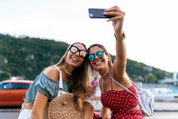 Happy young female friends in sunglasses sitting and taking a selfie on mobile phone  of city street