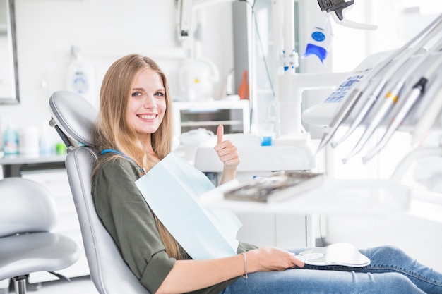 Happy young female patient sitting on dental chair gesturing ok sign