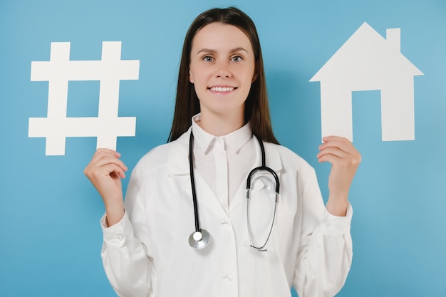 Happy young female physician practitioner in white uniform with stethoscope holds hashtag sign and house model, posing on blue background wall. Coronavirus, Quarantines and Social Distancing concept