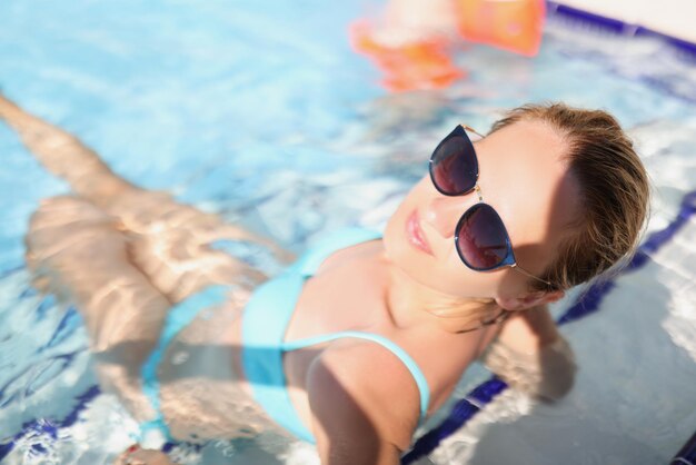 Happy young female sit in pool wearing trendy glasses