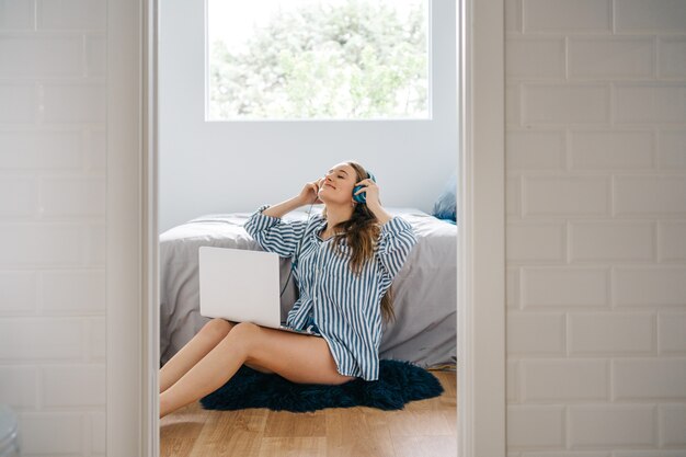 happy young female siting in her bedroom browsing on notebook while listening to music