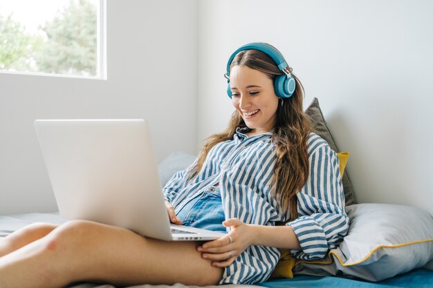 happy young female siting in her bedroom browsing on notebook while listening to music