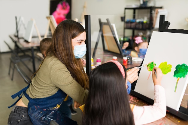 Happy young female teacher and an elementary girl with face masks are painting a landscape of trees on a canvas during an art class