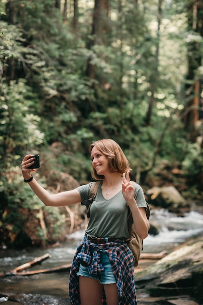 Happy young female traveler with backpack making selfie while standing by the mountain river