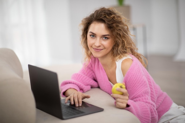 Happy young female using laptop and eating apple at home