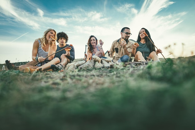 Happy young friends enjoy a sunny day at the mountain. They're laughing and grilling sausages on sticks over a campfire near tent.