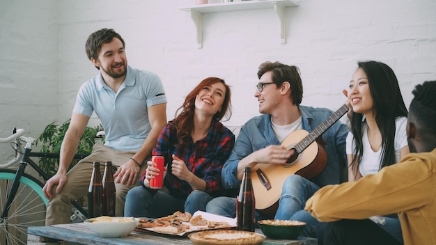 Happy young friends have party at shared apartment and singing together while their friend playing guitar at home