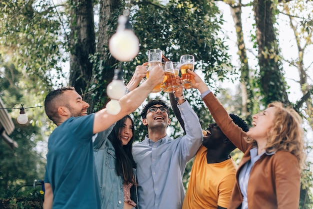 Happy young friends toasting with beer together cheerful people clinking glasses outdoor at party multiethnic group having fun and celebrating