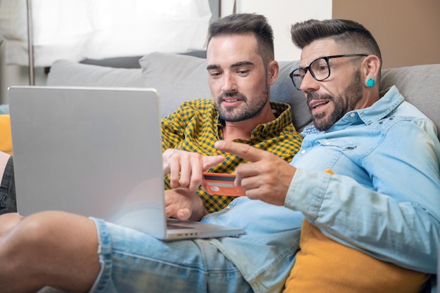 Happy young gay couple using laptop computer while sitting on a couch at home shopping online with a credit card