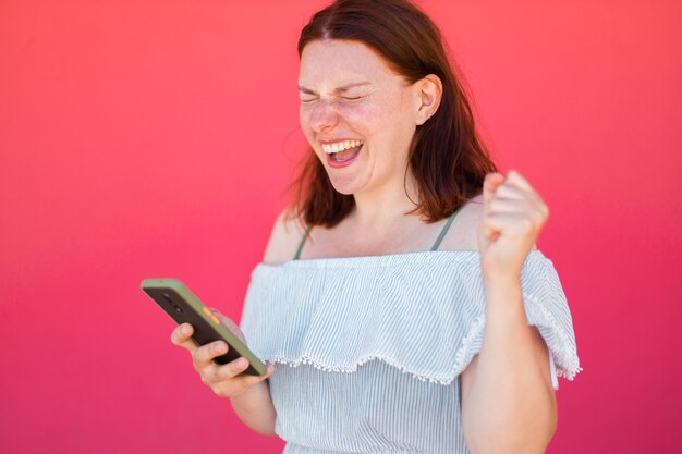 Happy young girl looking at smartphone screen and winning internet on pink wall, celebrating a victory