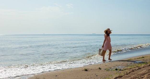 Happy young girl in a pink dress straw hat and with summer bag going on the waves of the sea at noon Relaxing unforgettable vacation at sea in summer Enjoying summertime Playing in water