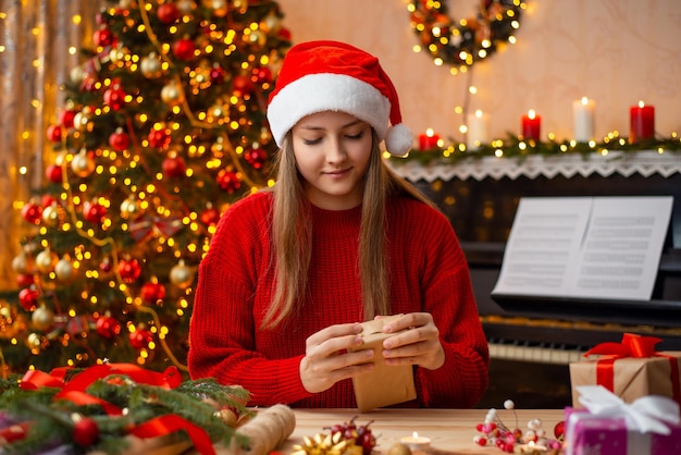 Happy young girl in Santa hat wrapping present boxes near beautifully decorated Christmas tree in living room Bright Christmas atmosphere celebrate holidays together with friends