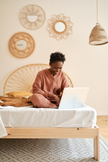 Happy young girl sitting on bed in bedroom and using laptop muslin pajama chatting with friends