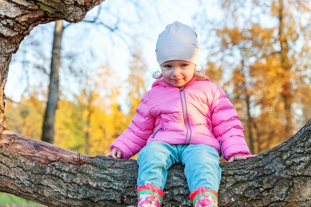 Happy young girl smiling and sitting on tree in beautiful autumn park on nature walks outdoors littl