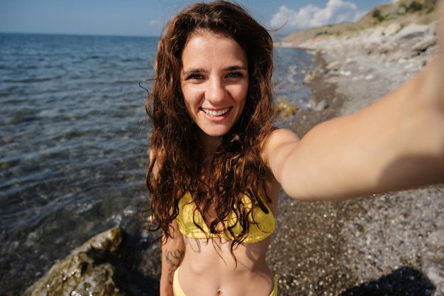 Happy young girl in a swimsuit takes a selfie on her phone on the beach against the background of the sea