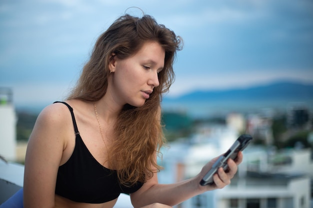 Happy young girl uses her phone on the roof of a house balcony city background a woman taking selfie at summer have a video chat smiling have fun photo of herself on mobile phone smartphone