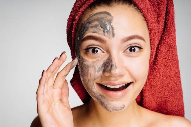 Happy young girl with a red towel on her head applied a useful clay mask to half the face, smiling