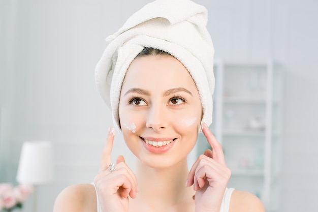 Happy young girl with a towel on her head apply a cleansing mask on her face