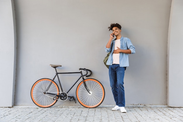 Happy young guy walking outdoors with bicycle talking by mobile phone