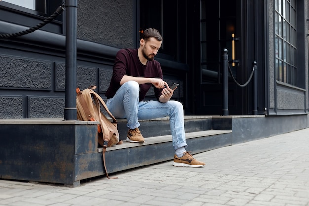 Happy young guy with beard relaxing with modern device outdoors. sitting on the steps and looking at a mobile phone