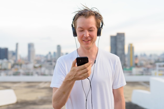 Happy young handsome man with blond hair using phone while listening to music against view of the city