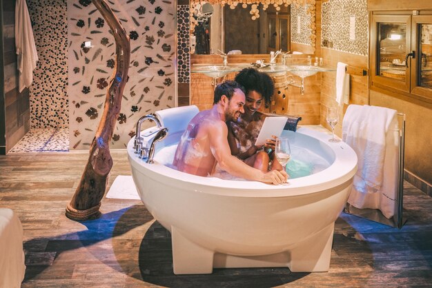 Happy young interracial couple relaxed enjoying their self in the jacuzzi drinking wine and with a tablet xA