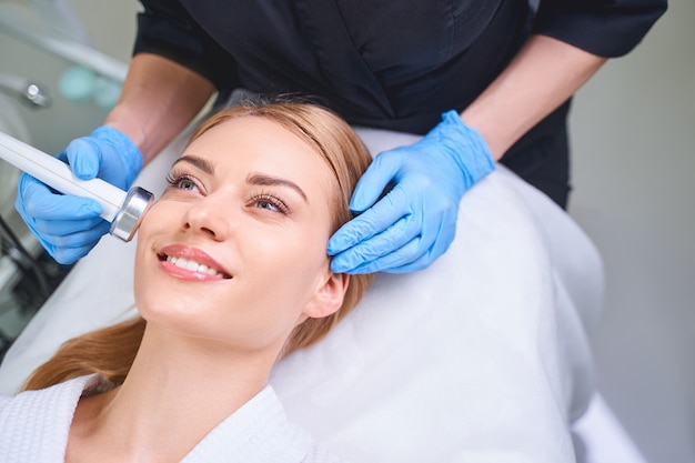 Happy young lady is taking care of her face by getting ultrasonic treatment by professional